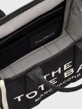 Bolso tipo tote de lona color negro con letras blancas bordadas en el frente que dicen 'THE TOTE BAG MARC JACOBS'. Cuenta con asas superiores cortas y una correa de hombro ajustable y extraíble.