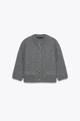 Chaqueta de punto grueso color gris, de cuello redondo y manga tres cuartos. Cierre frontal con botones joya dorados con detalles de pedrería. Cuenta con dos bolsillos frontales tipo parche.