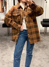 Camisa tipo sobrecamisa de corte oversize, confeccionada en tela a cuadros en tonos marrones y negros. Presenta cierre frontal con botones metálicos a presión, bolsillos frontales con solapa y mangas largas con puños abotonados.
