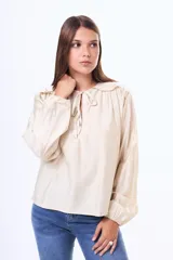 Blusa de satén color beige, con cuello redondo, detalle de volados en los hombros y cierre frontal con lazo. Presenta mangas largas con puños elásticos.