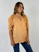 Blusa de lino color mostaza con escote en V y mangas cortas abullonadas con terminación de volados y flecos. Presenta un diseño de corte holgado y textura natural.