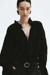 Blusa oversized color negro en tejido liviano de mezcla de lino y viscosa, con cuello elevado plisado y abertura en V con tiras finas. Mangas raglán extralargas con detalles bordados tono sobre tono y puños acampanados con pliegues.