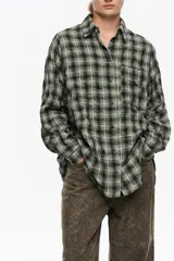 Camisa de corte recto y largo medio, confeccionada en mezcla de algodón con textura. Presenta un diseño de cuadros en tonos verde, negro y blanco, con cuello camisero, cierre frontal de botones, manga larga con puños abotonados y bolsillo de plastrón en el pecho.