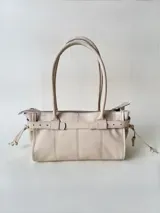 Cartera de cuero de tamaño medio con estructura rígida. Presenta una tira decorativa con tachas y hebillas metálicas en los extremos, además de lazos laterales. Cuenta con doble asa para llevar al hombro y cierre superior.