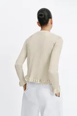 Chaqueta de punto canalé en color beige con hilo metalizado, cuello redondo, manga larga y cierre frontal con botones. Presenta detalles de volantes en la tapeta de los botones, el ruedo y los puños.
