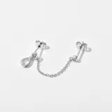 Aro doble plateado, unidos por una cadena de eslabones. Uno de los aros tiene un dije colgante con forma de lágrima y el otro un pequeño dije circular.