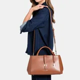 Cartera Guess modelo Carrie de color marrón, confeccionada en cuero sintético, con doble asa superior y correa de hombro ajustable. Presenta herrajes dorados, logo frontal de la marca y detalle de llavero colgante con candado.