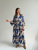 Vestido largo azul con estampado de hojas en tonos beige y blanco, escote en V con botones frontales, mangas largas amplias y diseño escalonado.
