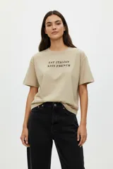 Remera de algodón color beige con cuello redondo y manga corta. Estampado frontal con texto en color marrón que dice "Eat Italian Kiss French".