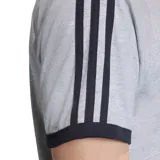 Remera de algodón gris con cuello y bordes de mangas en color negro. Presenta las icónicas tres tiras de Adidas en los hombros y el logo del trébol bordado en el pecho.