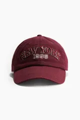 Gorra en sarga de algodón de color bordó, con visera curva, ojales bordados y el texto 'NEW YORK 1996' bordado en el frente.