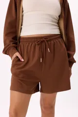Conjunto deportivo compuesto por un buzo con cierre frontal, capucha y bolsillos, y un short con cintura elástica y cordón ajustable. Confeccionado en tejido soft premium de color marrón.