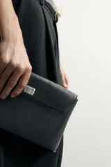 Bolso de mano tipo sobre negro, de acabado liso, con solapa y cierre de pieza metálica giratoria. Incluye asa de mano.