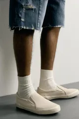 Zapatilla casual sin cordones de tipo slip-on, confeccionada en cuero con acabado de serraje y tejido en color beige. Cuenta con elásticos laterales en el empeine y suela de goma a tono.