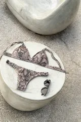 Bikini de dos piezas color marrón, con corpiño triangular fijo con corte bajo el busto, breteles regulables con hebilla y almohadillas desmontables. La bombacha es vedetina con frunce elástico en los costados.