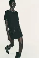 Top negro de manga corta con cuello redondo, adornado con perlas blancas aplicadas en el escote y en los puños de las mangas. Cierre posterior con botones de perla.