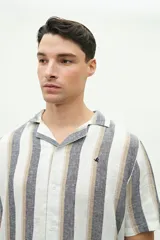 Camisa de manga corta con cuello cubano, confeccionada en una mezcla de lino y algodón con textura. Presenta un diseño a rayas verticales en tonos beige y celeste sobre fondo blanco, con cierre frontal de botones y logo bordado en el pecho.