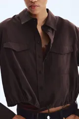 Camisa marrón de manga larga, corte corto y diseño holgado, con cuello clásico, cierre frontal de botones, bolsillos con solapa en el pecho y cordón ajustable en el bajo.