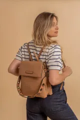 Mochila tipo cartera con tapa, confeccionada en símil cuero color beige. Presenta un diseño moderno con detalle de ojales metálicos en la parte frontal, compartimentos internos organizadores y bolsillo trasero con cierre.