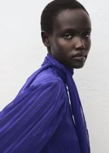 Blusa de manga larga en color azul eléctrico con diseño de rayas verticales sutiles. Presenta un cuello alto con detalle de lazo integrado y cierre frontal con botones ocultos. El tejido es semitransparente con acabado satinado.