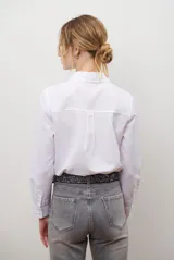 Camisa blanca de manga larga con cuello clásico y botones frontales. Tiene un bolsillo en el pecho con un pequeño logo bordado.