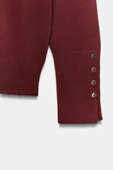 Jersey de punto liso en color beige, con cuello alto y manga larga con detalle de botones metálicos en los puños.