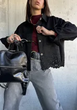 Chaqueta negra de gamuza con cuello camisero, cierre frontal de botones y detalle de bordado calado en el ruedo. Presenta mangas largas con puños abotonados y bolsillos frontales tipo parche.