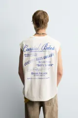 Camiseta tipo tank (sin mangas) de corte relajado, color blanco roto con efecto lavado. Presenta un estampado frontal en azul con texto en francés: "Coquillages Grillés De Côte-Marine" y "Salty Shack".
