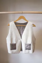 Chaleco reversible confeccionado en cuero genuino. Presenta un lado de piel de oveja gris con bolsillos de cuero negro y un lado de cuero liso gris. Diseño sin mangas con cierre frontal.