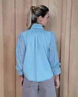Camisa de denim celeste con detalles de broderie en las mangas, cuello alto con volados y canesú con frunces. Presenta cierre frontal con botones y mangas largas con puños elásticos.