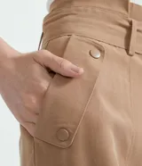 Pantalón cargo de tiro medio color beige, con cintura alta fija, cinturón ajustable para anudar, bolsillos laterales con solapa y botones, recortes verticales en las piernas y cierre frontal con botón y cremallera.