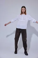 Camisa blanca de algodón con pechera decorada con alforzas y puntillas. Presenta cierre frontal con botones, mangas tres cuartos con frunces y detalles de puntilla, y un diseño de corte holgado con ruedo redondeado.