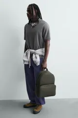 Mochila de color verde militar con textura grabada, diseño minimalista y forma redondeada. Cuenta con un compartimento principal y un bolsillo frontal, ambos con cierre de cremallera. Posee asa de mano y asas de hombro acolchadas y ajustables.