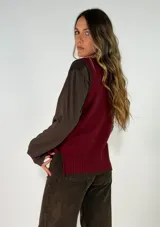 Chaleco de punto color marrón con detalles en color mostaza en el cuello, sisas y ruedo. Presenta un diseño de corte recto y un pequeño aplique decorativo en forma de flor en la parte inferior.