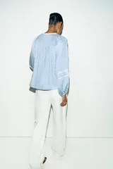 Blusa de rayas verticales blancas y celestes, con cuello redondo y escote en pico. Presenta detalles de bordados perforados y puntillas en el pecho y las mangas. Tiene mangas largas abullonadas con puños elásticos y cierre frontal con botones forrados.
