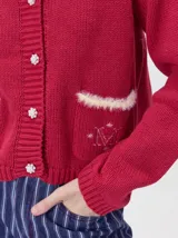 Cardigan de algodón grueso color rojo, con cuello redondo, cierre frontal de botones grandes y bolsillos cuadrados en el frente. Presenta detalles de pelo sintético en el cuello y los bolsillos, y logo bordado en uno de los bolsillos.
