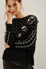 Sweater negro de punto con cuello redondo, manga larga y diseño holgado. Presenta aplicaciones de lentejuelas plateadas dispersas, bordes plateados en las mangas y apliques florales con lentejuelas en el hombro y frente.