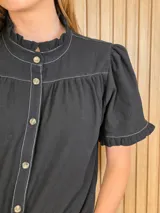 Camisa de manga corta color beige, con cuello alto con volados, botonadura frontal y mangas abullonadas con terminación fruncida.