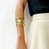 Brazalete ancho de bronce dorado, con textura martillada, compuesto por tres bandas unidas y adornado con tres piedras engarzadas: una central ovalada de nácar blanco y dos laterales cuadradas de nácar blanco.