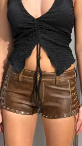 Short de cuero color marrón, tiro bajo, con tachas metálicas en el borde inferior y cordones entrelazados en los laterales.