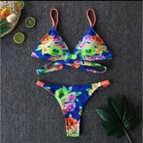 Bikini de dos piezas con estampado floral sobre fondo azul. El top es de estilo triángulo con tiras naranjas y diseño cruzado en la parte inferior del busto. La bombacha es tipo tanga con laterales regulables y detalles en color naranja.