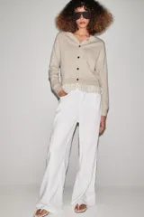 Chaqueta de punto color beige con cuello redondo, manga larga y cierre frontal con botones. Presenta un diseño combinado con volados de tela en el bajo.