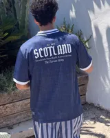 Camiseta de fútbol de Escocia de manga corta, color azul marino con diseño a cuadros sutiles, cuello en V y cuello camisero blanco con líneas azules. Presenta detalles estampados en el frente, incluyendo el escudo de la Asociación de Fútbol de Escocia, un león rampante y el texto 'Scottish Football Association'.