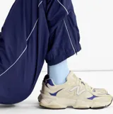 Championes urbanos New Balance modelo 9060, con diseño futurista inspirado en la estética de los años 2000. Presentan una combinación de tonos beige y blanco con detalles en azul, entresuela esculpida con tecnología de amortiguación ABZORB y SBS, y una capellada de materiales mixtos.