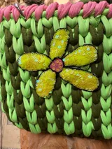Cartera tejida a crochet en hilo de algodón color verde con detalles en rosa y aplique de flor amarilla con lentejuelas. Tiene doble asa corta y correa larga de cadena metálica.