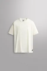 Camiseta deportiva de running color blanco crudo, confeccionada en tejido técnico ligero con microperforaciones. Presenta cuello redondo, manga corta y un pequeño detalle de logo estampado en negro en el bajo delantero.