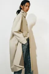 Gabardina larga de corte recto y diseño minimalista en color beige, con cuello solapa, manga larga y cierre frontal con botones. Cuenta con una capa desmontable en el mismo tejido y una solapa decorativa en el pecho.