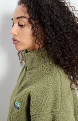 Campera de corderito con efecto shearling, color verde oliva, de corte holgado, cuello alto y cierre frontal completo. Presenta un pequeño parche con logo en el pecho.