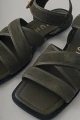 Sandalia plana de cuero negro, con diseño minimalista de tiras finas cruzadas sobre el empeine y una tira ajustable con hebilla metálica en el tobillo.
