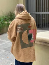 Campera tipo hoodie confeccionada en tejido de corderito color camel, con diseño de collage en la espalda que combina parches de diferentes texturas, colores y estampados geométricos. Presenta capucha y corte holgado.